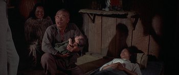 Movie still from “The Hawaiians” (1970), directed by Tom Gries – A man holding a baby in his arms while a woman lays on the ground; Medium shot, High angle