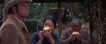 Movie still from “The Hawaiians” (1970), directed by Tom Gries – A woman eating a piece of bread while sitting next to a man; Medium shot, Over the shoulder angle