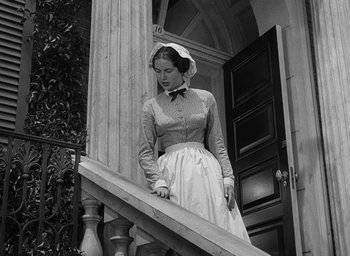 Movie still from “The Heiress” (1949), directed by William Wyler – A woman in a white dress and a white hat standing on a staircase; Medium shot, Low angle