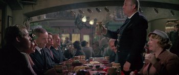 Movie still from “The Hindenburg” (1975), directed by Robert Wise – A man is holding a jar in front of a group of diners; Medium shot, Over the shoulder angle