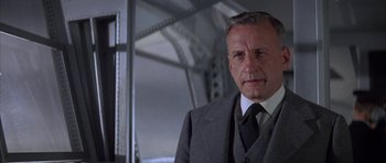 Movie still from “The Hindenburg” (1975), directed by Robert Wise – A man in a suit and tie standing in front of a bridge; Close Up shot, Low angle