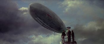 Movie still from “The Hindenburg” (1975), directed by Robert Wise – Three men are standing on a platform looking at an airship in the sky; Extreme Wide shot, Low angle