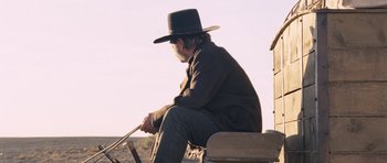 Movie still from “The Homesman” (2014), directed by Tommy Lee Jones – An older man sitting on a bench with a hat on; Medium shot, Over the shoulder angle