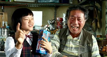 Movie still from “The Host” (2006), directed by Bong Joon Ho – An older man and a young girl are smiling for the camera; Medium shot, Over the shoulder angle