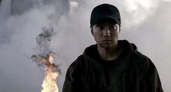 Movie still from “The Host” (2006), directed by Bong Joon Ho – A man wearing a baseball cap standing in front of a fire; Close Up shot, Low angle
