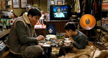 Movie still from “The Host” (2006), directed by Bong Joon Ho – A man and a boy eating food at a table; Medium shot, High angle