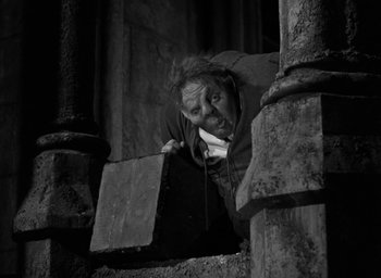 Movie still from “The Hunchback of Notre Dame” (1939), directed by William Dieterle – A man leaning on a wooden post looking up at the ground; Wide shot, Low angle