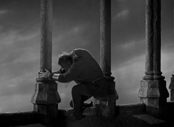 Movie still from “The Hunchback of Notre Dame” (1939), directed by William Dieterle – A person kneeling down on the ground in front of pillars; Wide shot, Low angle