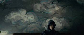 Movie still from “The Imposter” (2012), directed by Bart Layton – A man in a hooded jacket sitting in front of a painting; Medium shot, Low angle
