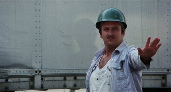 Movie still from “The In-Laws” (1979), directed by Arthur Hiller – A man in a hard hat and overalls standing in front of a truck; Medium shot, Low angle