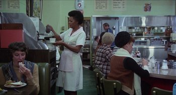 Movie still from “The In-Laws” (1979), directed by Arthur Hiller – A woman in a white apron in front of some people; Medium shot, Over the shoulder angle