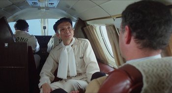 Movie still from “The In-Laws” (1979), directed by Arthur Hiller – An older man sitting on a plane talking to another man; Medium shot, Over the shoulder angle