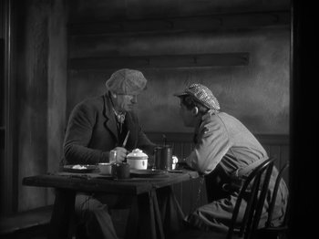 Movie still from “The Informer” (1935), directed by John Ford – Two men are sitting at a table and one of the men is smoking a cigarette; Medium shot, High angle