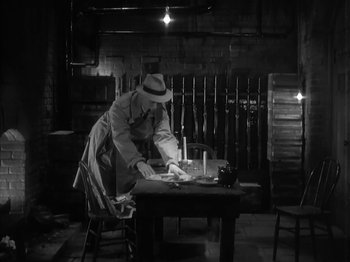 Movie still from “The Informer” (1935), directed by John Ford – A man in a hat and a trench coat standing at a wooden table; Medium shot, High angle