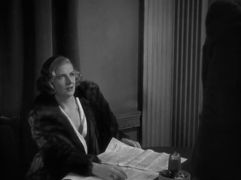 Movie still from “The Informer” (1935), directed by John Ford – A woman sitting at a table with papers in front of her; Medium shot, Over the shoulder angle