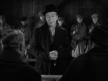 Movie still from “The Informer” (1935), directed by John Ford – An older man in a hat and a coat is holding a pen in front of a group of people; Medium shot, Over the shoulder angle