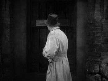 Movie still from “The Informer” (1935), directed by John Ford – A man in a trench coat and a hat standing in front of a wooden door; Medium shot, Over the shoulder angle