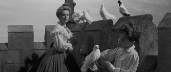 Movie still from “The Innocents” (1961), directed by Jack Clayton – A man and a woman standing in front of a flock of pigeons; Medium shot, Low angle