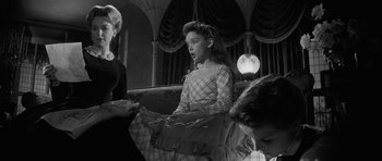 Movie still from “The Innocents” (1961), directed by Jack Clayton – A young girl is sitting on a couch while two other girls watch; Medium shot, Over the shoulder angle