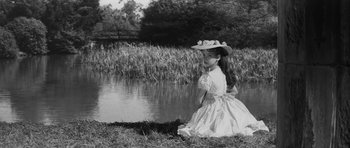 Movie still from “The Innocents” (1961), directed by Jack Clayton – A young girl in a dress and a hat by a pond; Medium shot, High angle