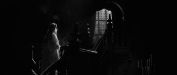 Movie still from “The Innocents” (1961), directed by Jack Clayton – A woman standing on top of a set of stairs next to a window; Wide shot, Over the shoulder angle