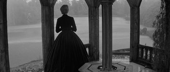 Movie still from “The Innocents” (1961), directed by Jack Clayton – A woman in a black dress standing next to a pillar; Wide shot, High angle