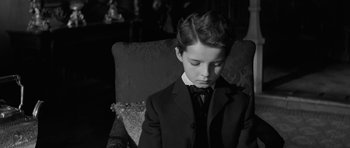 Movie still from “The Innocents” (1961), directed by Jack Clayton – A young boy in a suit and bow tie sitting in a chair; Medium shot, High angle