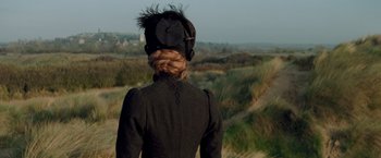 Movie still from “The Invisible Woman” (2013), directed by Ralph Fiennes – A woman in a black coat and a black hat looking out over a field; Medium shot, Over the shoulder angle