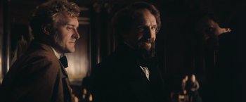 Movie still from “The Invisible Woman” (2013), directed by Ralph Fiennes – A man with a beard standing next to another man in a suit; Medium shot, Over the shoulder angle