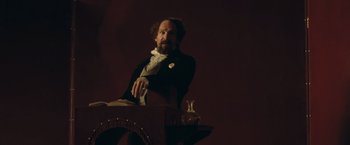 Movie still from “The Invisible Woman” (2013), directed by Ralph Fiennes – A man sitting at a table in a dark room; Medium shot, Low angle