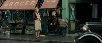 Movie still from “The Iron Lady” (2011), directed by Phyllida Lloyd – A woman standing in front of a store with a broom; Wide shot, Over the shoulder angle