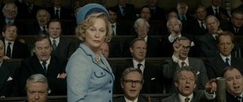 Movie still from “The Iron Lady” (2011), directed by Phyllida Lloyd – A woman in a blue suit standing in front of a group of people; Medium shot, Low angle