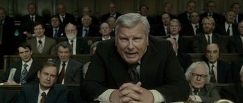 Movie still from “The Iron Lady” (2011), directed by Phyllida Lloyd – An older man sitting in front of a group of people; Medium shot, Over the shoulder angle