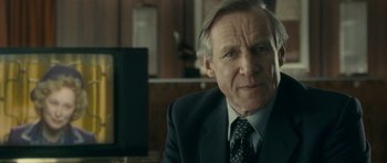 Movie still from “The Iron Lady” (2011), directed by Phyllida Lloyd – An older man wearing a suit and tie in a room; Close Up shot, Over the shoulder angle