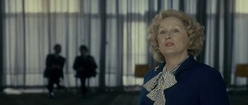 Movie still from “The Iron Lady” (2011), directed by Phyllida Lloyd – An older woman in a blue dress and a man in a black suit in the background; Medium shot, Low angle