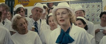 Movie still from “The Iron Lady” (2011), directed by Phyllida Lloyd – An older woman wearing a white hat standing in front of a group of people; Medium shot, Over the shoulder angle