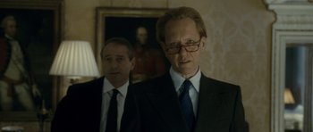 Movie still from “The Iron Lady” (2011), directed by Phyllida Lloyd – A man in a suit and tie standing next to another man in a suit and tie; Medium shot, Over the shoulder angle