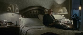 Movie still from “The Iron Lady” (2011), directed by Phyllida Lloyd – An older man sitting on top of a bed reading a magazine; Medium shot, Low angle
