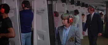 Movie still from “The Island” (1980), directed by Michael Ritchie – A young man wearing ear muffs while standing in front of an audience; Medium shot, Over the shoulder angle