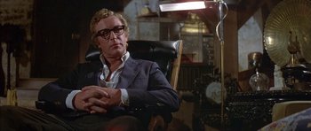 Movie still from “The Island” (1980), directed by Michael Ritchie – A man sitting in a chair wearing a suit and glasses; Medium shot, Low angle