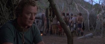 Movie still from “The Island” (1980), directed by Michael Ritchie – A man sitting next to a tree in front of a group of people; Medium shot, Over the shoulder angle