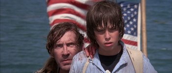 Movie still from “The Island” (1980), directed by Michael Ritchie – A man and a boy are standing next to each other in front of an american flag; Close Up shot, Low angle