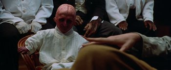 Movie still from “The Island of Dr. Moreau” (1996), directed by Richard Stanley – A man with a bald head sitting next to other people; Close Up shot, High angle