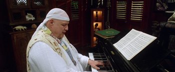 Movie still from “The Island of Dr. Moreau” (1996), directed by Richard Stanley – An older man playing a piano in front of a book case; Medium shot, High angle