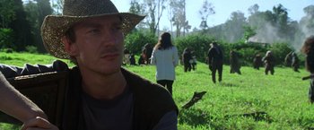 Movie still from “The Island of Dr. Moreau” (1996), directed by Richard Stanley – A man wearing a straw hat in a grassy field with a group of people; Close Up shot, Over the shoulder angle