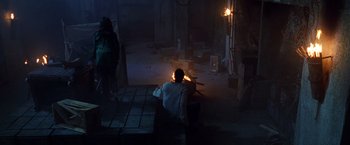 Movie still from “The Island of Dr. Moreau” (1996), directed by Richard Stanley – A man sitting on top of a bench next to a fire pit; Wide shot, High angle