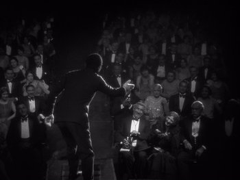 Movie still from “The Jazz Singer” (1927), directed by Alan Crosland – A man in a suit and tie holding a microphone in front of an audience; Wide shot, High angle