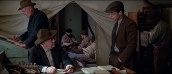 Movie still from “The Journey of Natty Gann” (1985), directed by Jeremy Kagan – A group of men sitting around a table in front of a tent; Medium shot, Over the shoulder angle