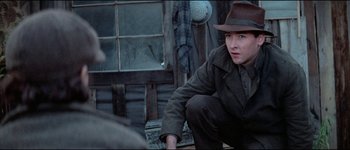Movie still from “The Journey of Natty Gann” (1985), directed by Jeremy Kagan – A man in a hat and a jacket sitting in front of a building; Medium shot, Over the shoulder angle