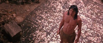 Movie still from “The Jungle Book” (1994), directed by Stephen Sommers – A shirtless man standing on the street with a bunch of coins; Wide shot, High angle